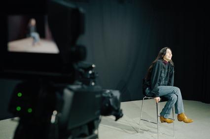 A person sits on a chair in front of a camera against a dark backdrop.