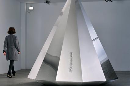 A person observing a large, metallic pyramid structure in a modern art gallery.