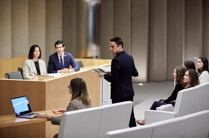A man speaks at a podium in a modern classroom setting while a group of attentive students and instructors listen.