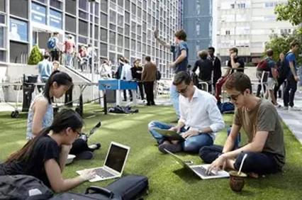 A group of students working on laptops outdoors in a lively urban setting.