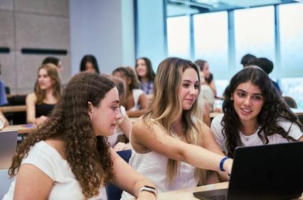 Three students collaborate on a laptop in a classroom filled with peers.