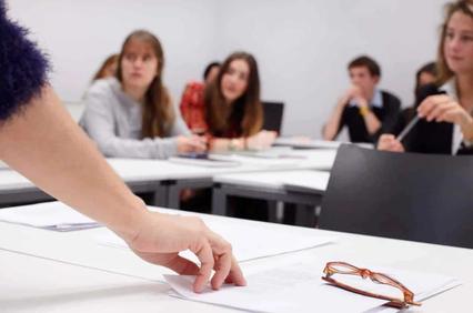 A classroom scene where a group of students are focused on a teacher pointing at papers on a table.