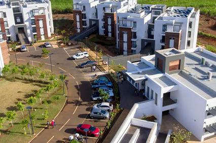 Aerial view of a modern housing complex with white buildings, parking lot, and a background of green fields and mountains.