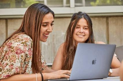 Two women smiling and looking at a laptop outdoors.