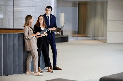 Three business professionals, two women and one man, are engaged in a discussion in a modern office lobby.