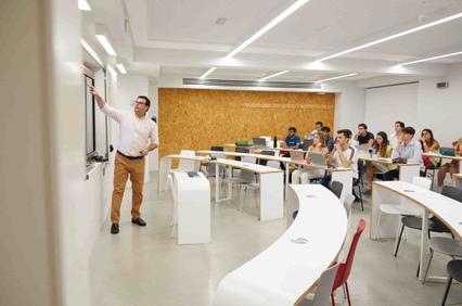 A teacher is explaining in a modern classroom with students seated at curved desks.
