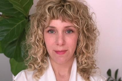 Portrait of a woman with curly blonde hair and a white shirt, standing in front of a green plant.