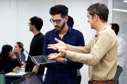 A man explains something to another man using a laptop in a classroom setting.