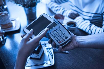 A person making a payment using a mobile phone, with a card reader held by another hand, over a cafe table with desserts.