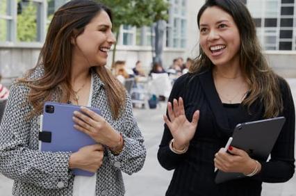 Two women are smiling and engaged in a friendly conversation outdoors.