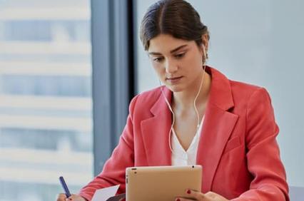 A woman in a red blazer is sitting at a table, working intently on a tablet while listening to music.