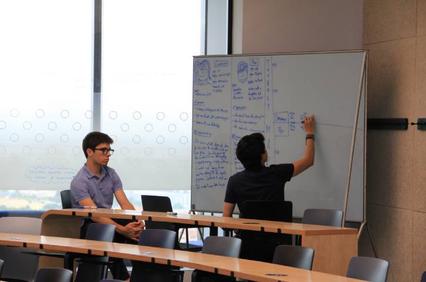 A student is writing on a whiteboard while another student sits and observes in a classroom.