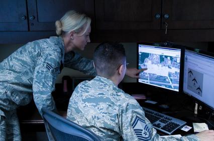Two military personnel, one female and one male, are analyzing satellite imagery on computer monitors in a dimly lit room.