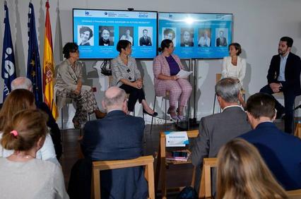 A panel discussion taking place in front of an audience, with digital banners displaying profiles of speakers in the background.