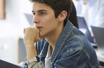 A young man in a denim jacket sits focused at a computer in a classroom setting, surrounded by other students also working on laptops.