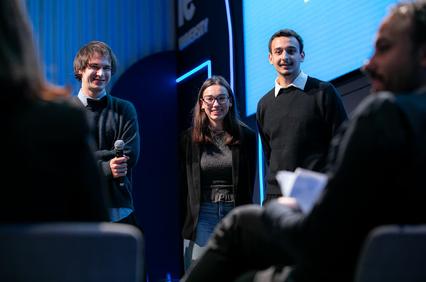 Three young individuals are standing on stage, engaging with an audience.