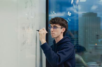 A young man is writing on a glass whiteboard in a modern office environment.