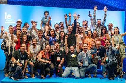 A group of people celebrating at a conference event with raised hands and cheerful expressions in front of a stage with 'IE University' branding.