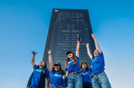 A group of joyful people in blue shirts celebrating in front of a tall modern building under a clear blue sky.