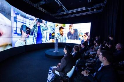 A group of people watching a large curved screen displaying various technology-related images.