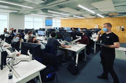 Office environment showing several people working at desks with computers, one man stands wearing a face mask.