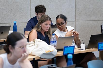 Two students are engaged in discussion at a classroom desk while others work on laptops.