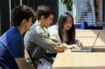 Three students are engaged in a discussion while looking at a laptop in a bright modern workspace.