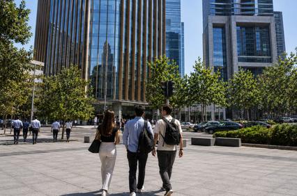 A group of people walking on a sidewalk in a modern urban area with skyscrapers in the background.