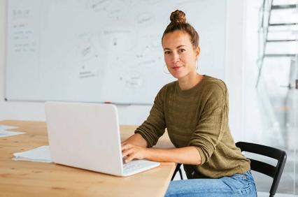 A woman sitting at a desk in an office, working on a laptop with a whiteboard in the background.