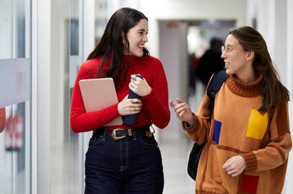Two women walking and talking in a corridor, one holding a laptop and the other holding coffee cups.