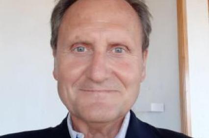 Portrait of a smiling elderly man in a suit jacket standing indoors.