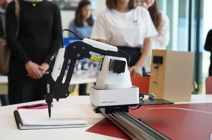 A robotic arm is positioned over a sheet of paper on a table, alongside an audience observing.