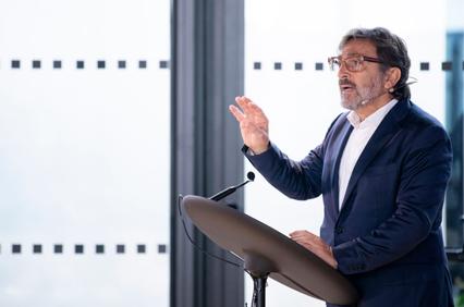 A man in a suit with glasses is speaking at a podium in a modern office environment.