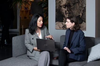 Two professional women are sitting on a couch, discussing a project on a laptop.