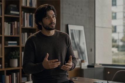 A man with dark hair is speaking passionately while gesturing in an office setting.