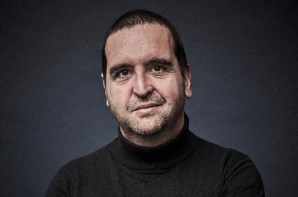 Portrait of a smiling man wearing a black turtleneck, set against a blue-grey background.