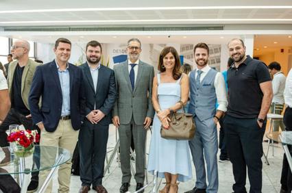A group of six adults, three men and three women, pose for a photograph at an indoor event with other attendees in the background.