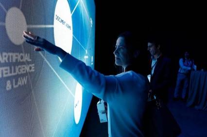 A woman interacts with a digital touchscreen display at a technology conference, with a male observer in the background.