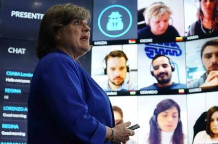 A woman in a blue jacket stands speaking in front of a large screen displaying multiple people in a virtual meeting.
