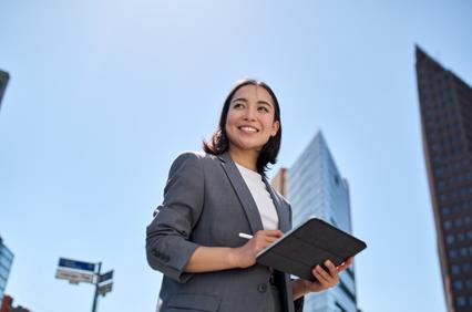 A businesswoman holding a tablet, smiling against a backdrop of modern skyscrapers under a clear blue sky.