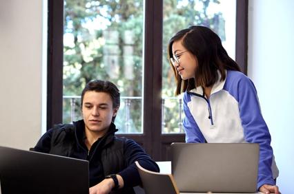 Two young adults, a male and a female, are using laptops and discussing something in a brightly lit room with windows showing trees outside.