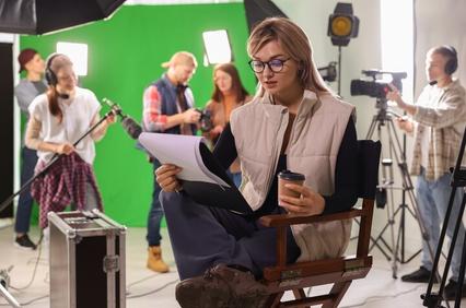 A woman sitting in a director's chair reading a script on a film set with crew members and equipment in the background.