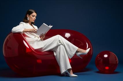A woman in a white suit sits elegantly on a red, sculptural seat while reading a book.