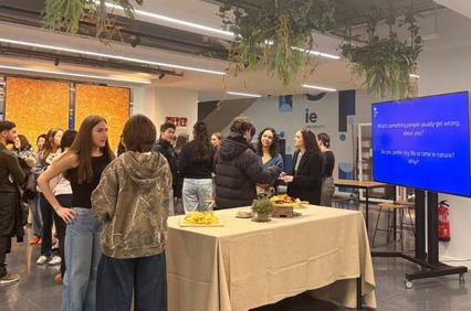 A lively gathering where people are engaged in discussion around a table with food and a presentation screen.