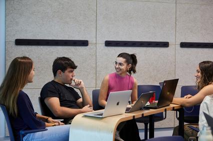 Four young adults are engaged in a discussion at a table with laptops in a modern classroom setting.