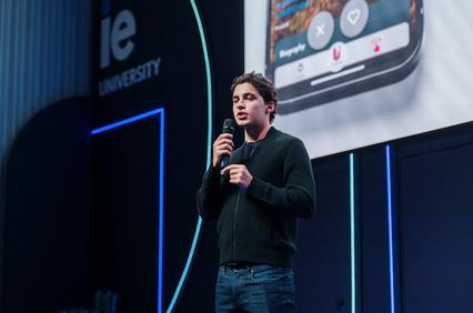 A speaker presents on stage at IE University, focusing on a mobile device.
