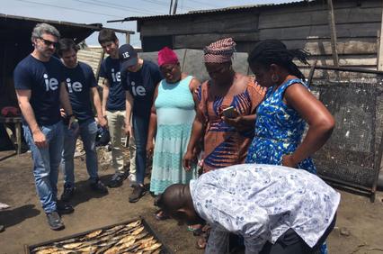 A group of people, including visitors wearing matching shirts, are shown observing local activities in a rustic outdoor setting.
