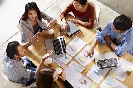 A group of five diverse professionals engaging in a meeting with laptops and documents on a wooden table.