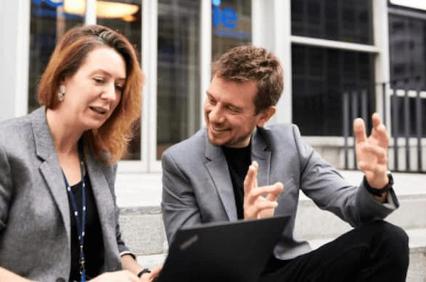 A man and woman, both in business attire, are sitting and looking at a laptop outdoors, with the man explaining something and gesturing with his hands.