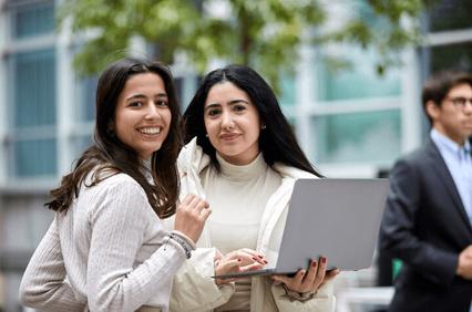 Two women holding a laptop and smiling outside a modern building.